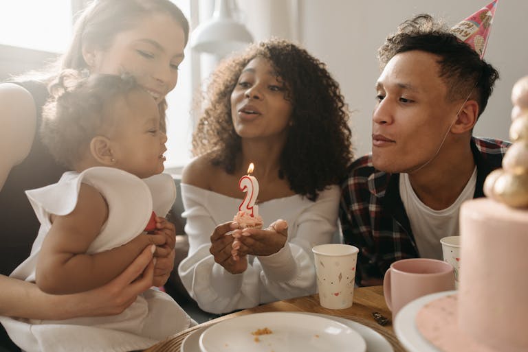 A diverse family celebrates a toddler's second birthday with a cupcake and candle indoors.